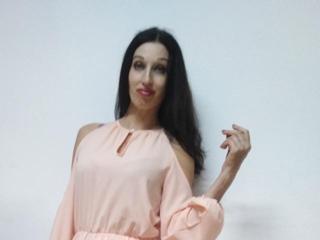 Jillian Pattie, com longos cabelos pretos, blusa rosa com um ombro à mostra, um sorriso adorável e a mão direita levantada, exibindo dedos finos. Sua blusa rosa tem um pequeno detalhe em formato de chave..