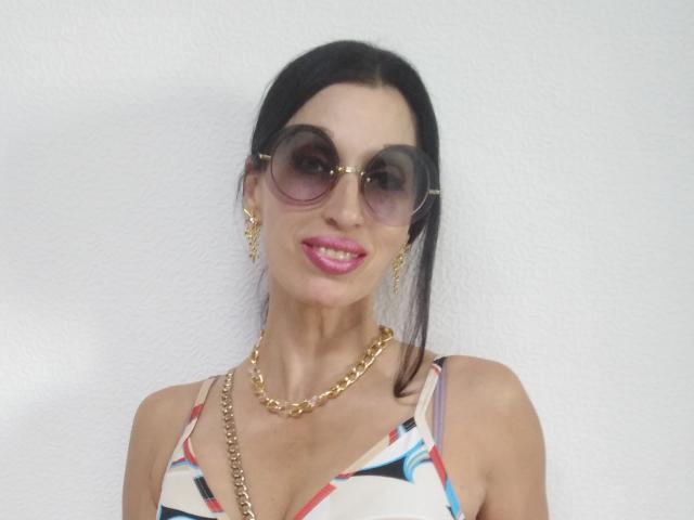 Jillian Pattie mit dunklem Haar, rosafarbenem Lippenstift, einer goldenen Kette, goldenen Ohrringen und großen, runden Sonnenbrillen, trägt ein farbenfrohes Oberteil mit Muster und lächelt..