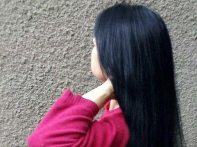 JaneLaLucky with long black hair, wearing a red sweater, touching her neck, standing against a textured brown wall.
