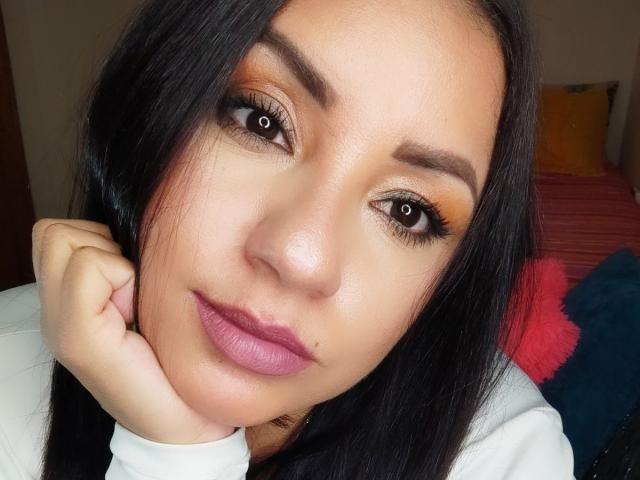 O rosto de CamilaBjQueen, pele bronzeada, cabelo preto longo, maquiagem nos olhos em tons de laranja e marrom, olhos castanhos, batom rosa, mão apoiada na bochecha, blusa branca, sorriso suave, leve brilho na maquiagem..