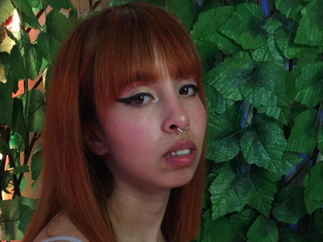 Samantha Morgan hat rotes Haar, schwarzen Eyeliner, rosigen Rouge, eine Nasenring und glänzende, rosafarbene Lippen, die leicht zu einem Mund formt sind, und sie blickt zur Seite. Im Hintergrund ist ein grüner, blattiger Hintergrund zu sehen..
