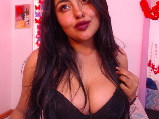 Angela Roggers, con cabello negro largo, lápiz labial morado oscuro y senos grandes cubiertos de encaje negro, sonriendo a la cámara, con la mano cerca del pecho. Habitación rosa con decoraciones rojas..