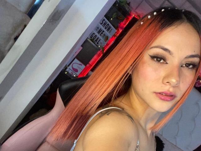 ValeryHarllow with orange-dyed hair, wearing a headband with small pearls, looking at the camera with pouty lips, showing off her tits in a pink chair.