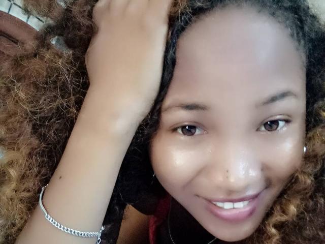 SaminahX, con cabello castaño rizado, sosteniendo una trenza, sonriendo con una piel brillante, ojos marrones, una nariz pequeña, vistiendo una pulsera de plata y pintalabios rosa..