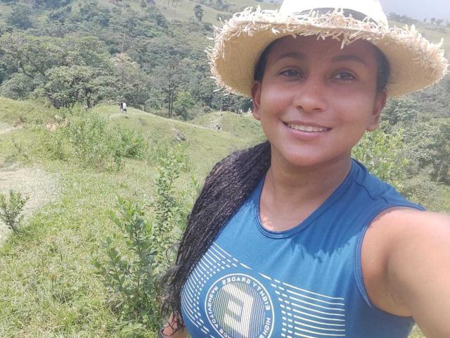 YiyaHot69 with tan skin, black braids, wearing a straw hat and blue sleeveless top, smiling in a grassy, hilly, forested area. Her ass is not visible.