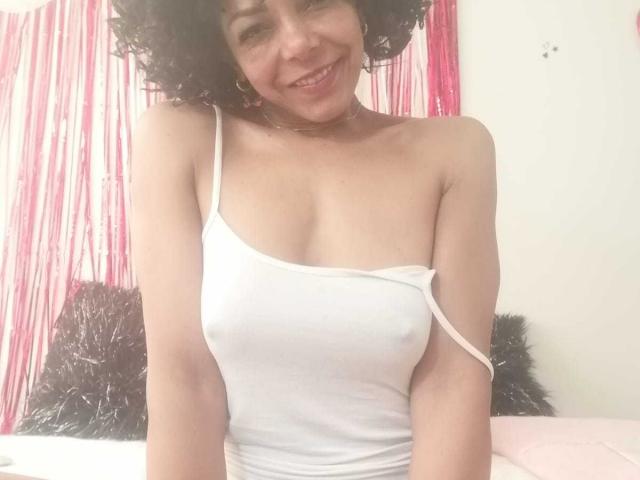 VenusChaud with curly dark hair, smiling, wearing a white tank top that shows her small, perky tits and nipples. Background has pink strings and black pillows.