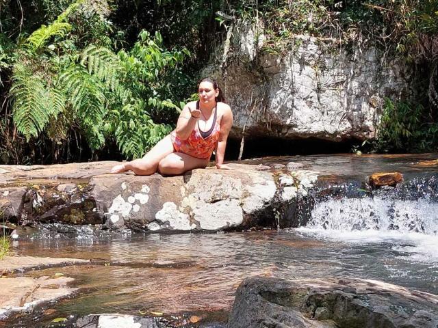 LadySelene with big ass, large boobs, and a red floral bikini, sitting on a rock, eating a banana, near a small waterfall in a lush, green jungle.