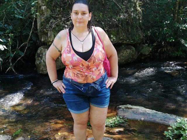 LadySelene has big tits, a tattoo on her right arm, and is wearing a pink floral top with blue denim shorts, standing by a rocky stream.