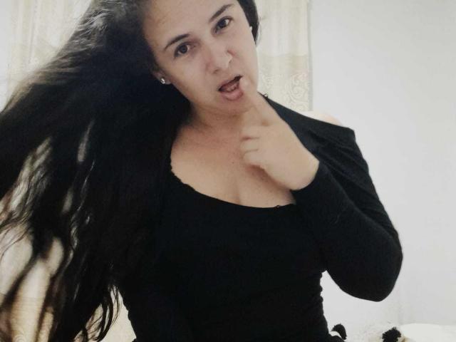 Brihana Charlot, con i suoi lunghi capelli neri, indossa una maglietta nera e ha un dito vicino alla bocca leggermente aperta, appare sorpresa o pensierosa. Il seno è visibile sotto la maglietta..
