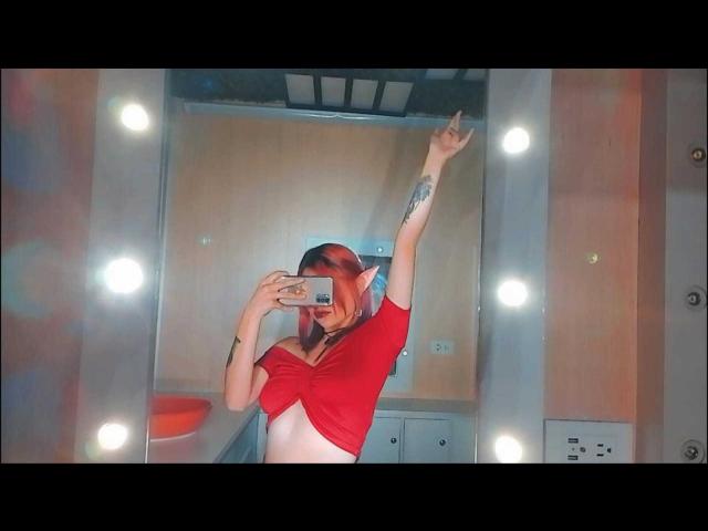 CameroonSweet, con cabello rojo, levantando su brazo derecho, apuntando hacia arriba, vistiendo una blusa roja, mostrando sus pequeños senos, sosteniendo un teléfono frente a un espejo..