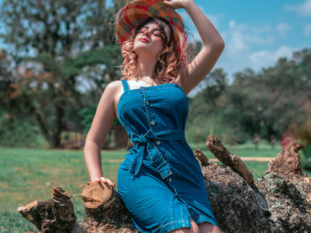 MaraMaison, com cabelo rosa ondulado, vestindo um vestido de jeans azul, segurando um chapéu colorido, sentada em um toco de árvore, com os seios à mostra, em um parque ensolarado..