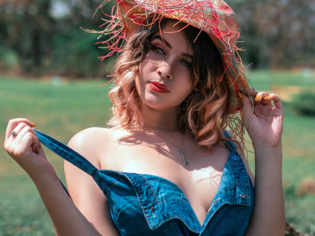 MaraMaison mit lockigem, rosa-braunem Haar, die einen roten Strohhut und ein blaues Jeanskleid trägt, zieht das Kleid herunter, um ihre Brüste zu zeigen, in einer sonnigen, offenen Umgebung..