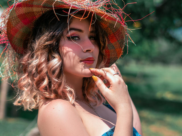 MaraMaison, avec ses cheveux blonds ondulés, portant un chapeau de paille coloré, touchant sa joue avec son doigt dont les ongles sont peints en orange, portant un haut à bretelles bleues et une bague à la main, souriant..