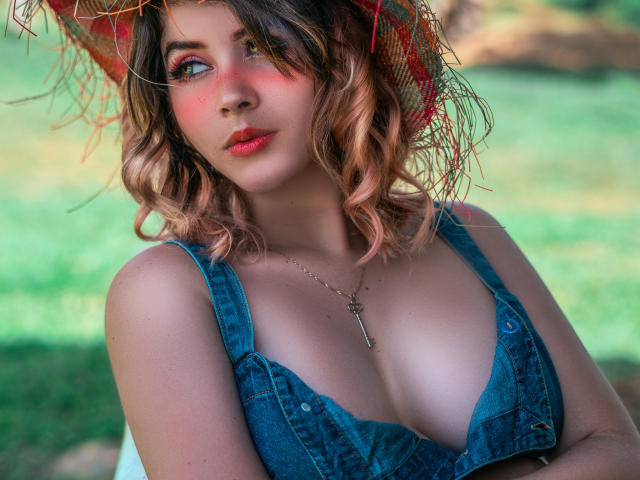 MaraMaison, con i capelli biondi ondulati, il rossetto rosso e l'abito di jeans blu, che mostra il seno, indossa un cappello di paglia rosso e una collana con una chiave, e guarda di lato..