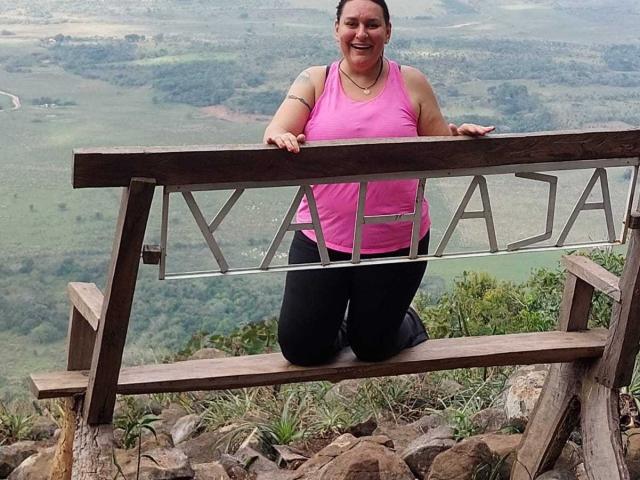 LadySelene, with medium tits and a pink tank top, kneels on a wooden bench with metal letters, smiling at the camera, wearing black pants, on a rocky hill.