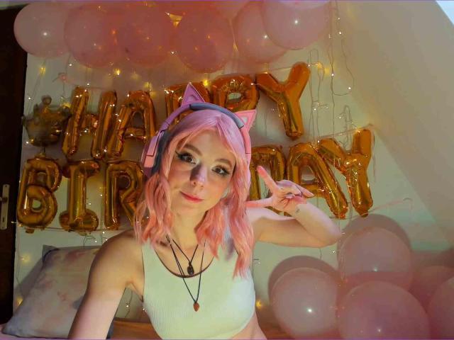 MissAstravert met roze haar, kattenoren en een koptelefoon, draagt een witte tanktop, maakt een vrede-teken en is omringd door roze ballonnen en gouden ballonnen met de tekst "Happy Birthday"..