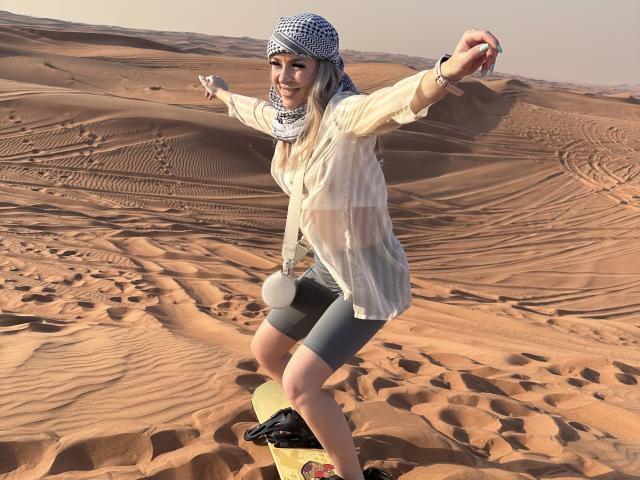 SophieDeee with a blue checkered headscarf, white shirt, gray shorts, and a yellow surfboard, standing in a desert, arms raised, smiling, in a sunny, sandy landscape.