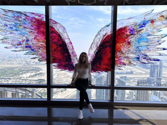 SophieDeee with colorful red, blue, and purple wings, sitting on a windowsill, wearing a white top, black pants, and white sneakers, in a high-rise building with a city view.