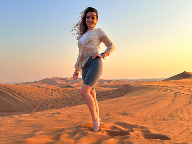 SophieDeee with long brown hair, wearing a sheer white blouse and tight blue shorts, standing in a sunlit desert, her legs and ass slightly angled, smiling at the camera.