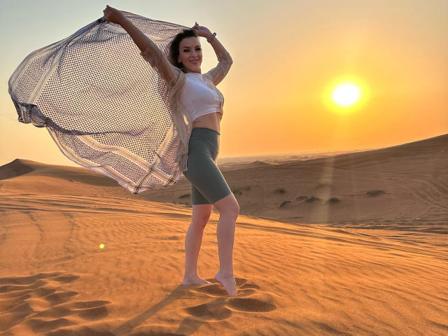 SophieDeee stands in a desert, arms raised, holding a mesh shirt, wearing a white crop top and green shorts, sun setting, barefoot, sand around her, smiling.