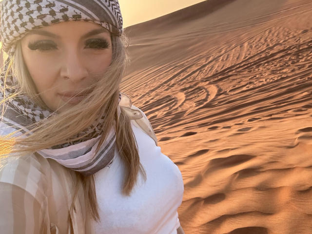 SophieDeee with long blonde hair, wearing a white shirt, checkered headscarf, and striped scarf, in a sunlit desert, showing her small tits and a serious face.