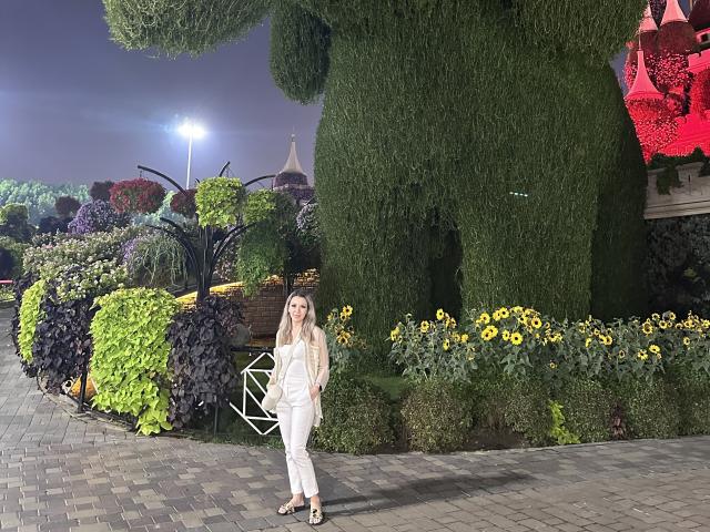 SophieDeee stands in front of a giant topiary, wearing a white outfit and black heels, with her tits modestly covered, in a nighttime garden with bright lights.