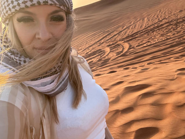 SophieDeee with a checkered headscarf, white shirt, and striped shawl, has blonde hair blowing in a desert sunset, showing her tits slightly against the orange sand dunes.