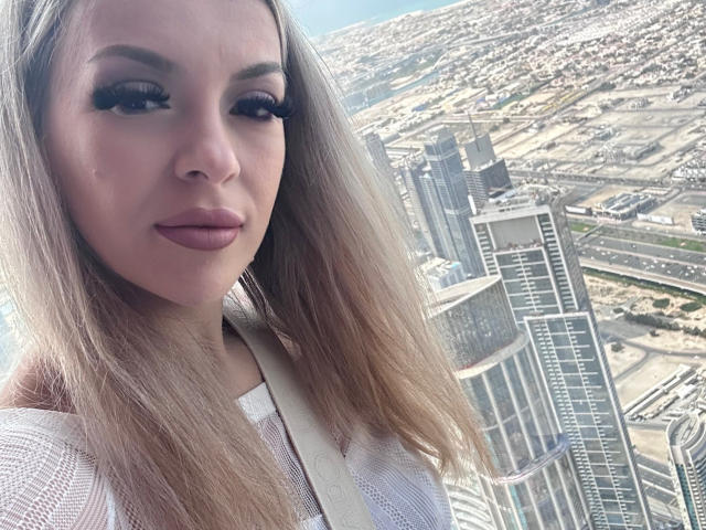 SophieDeee, com cabelos loiros longos, lábios cheios e cílios grossos, vestindo uma blusa branca de tela, está em pé perto de um horizonte urbano com edifícios altos. Seios não visíveis..