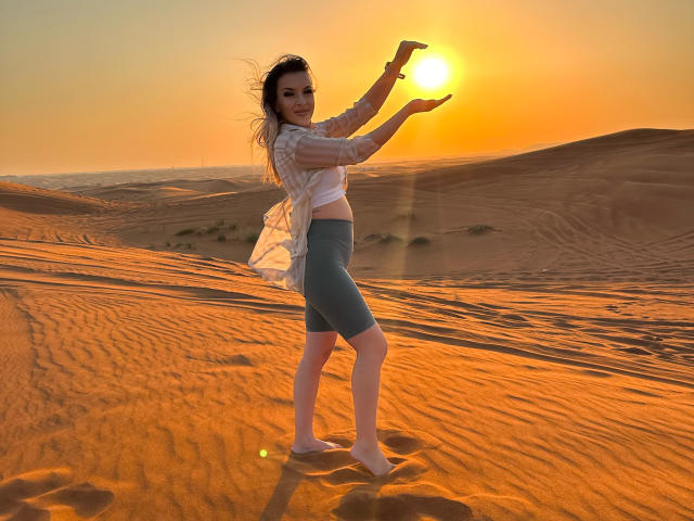 SophieDeee with sun in background, arms raised, wearing white top, blue skirt, standing barefoot in orange desert, sun casting glow, wind blowing through her hair, serene expression.