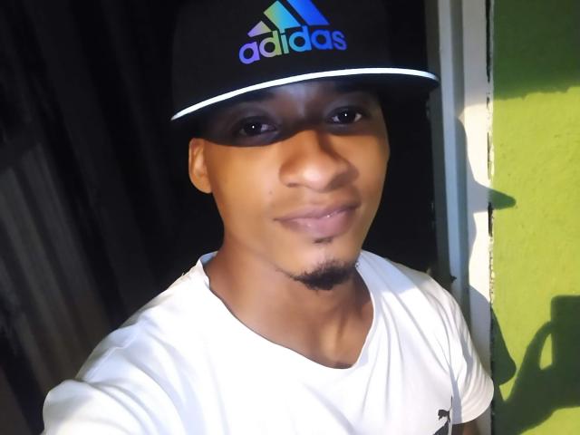 TheDon69, com pele escura, pequeno bigode, usando um boné preto da Adidas e uma camiseta branca, sorrindo suavemente, com sombra no lado direito..