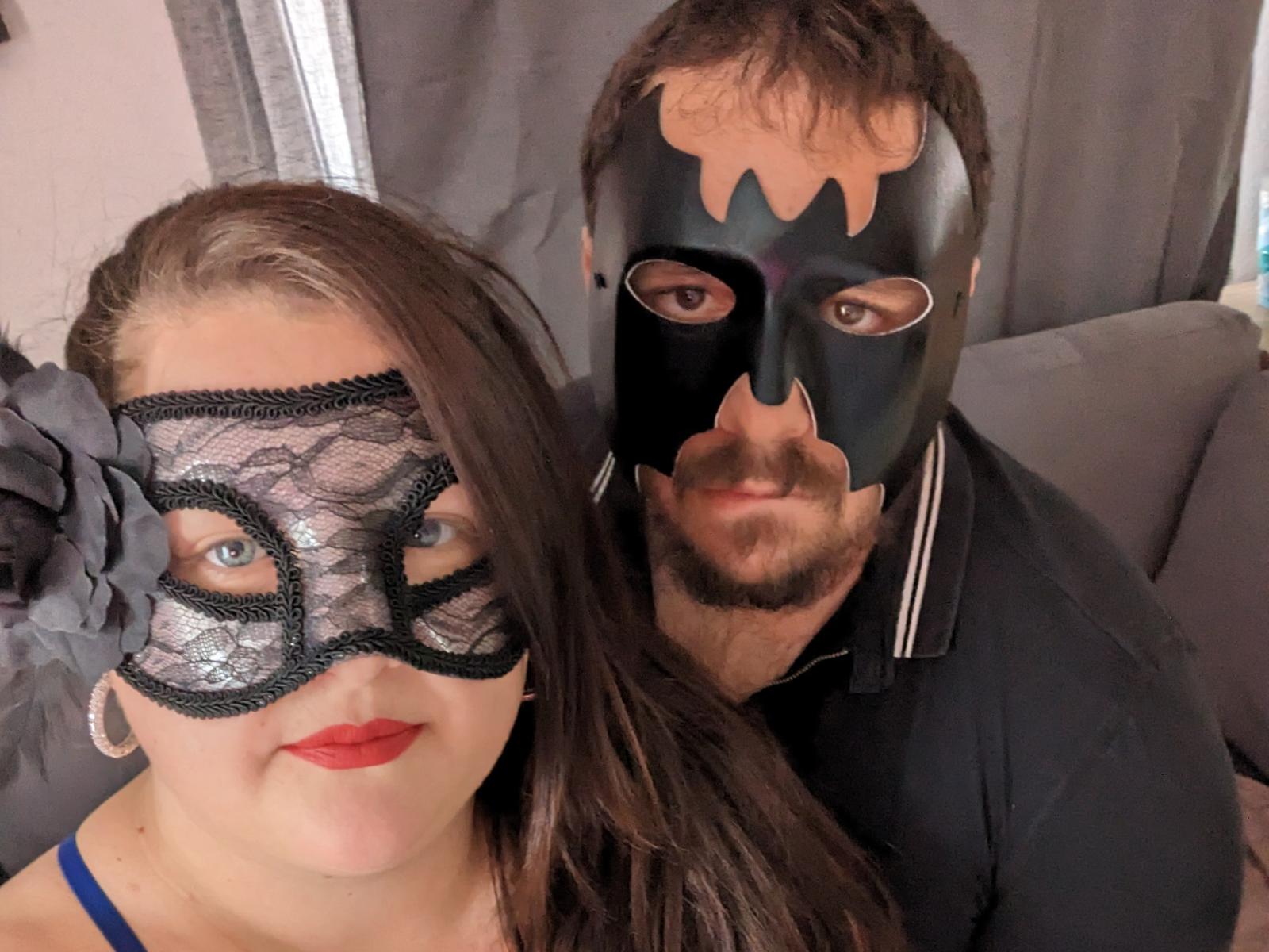 XCoupleMasqueX con ojos azules, lápiz labial rojo, máscara de encaje negro y cabello castaño largo. XCoupleMasqueX, con barba, en máscara negra, camisa negra, de pie detrás. Ambos sonriendo..