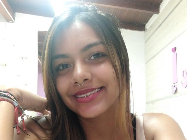 Emily Roberts tem a pele clara, cabelos castanhos longos, um piercing no nariz e está sorrindo, mostrando seus aparelhos ortodônticos de metal. Ela está usando batom rosa e pulseiras vermelhas..