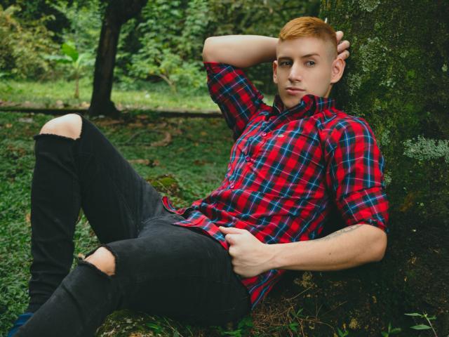 Jonathan Jeremiah, met een rood geruit shirt, zwarte versleten jeans en kort oranje haar, leunt tegen een mosgroene boom, rechterhand achter zijn hoofd, linkerhand op zijn knie, met een serieuze blik..