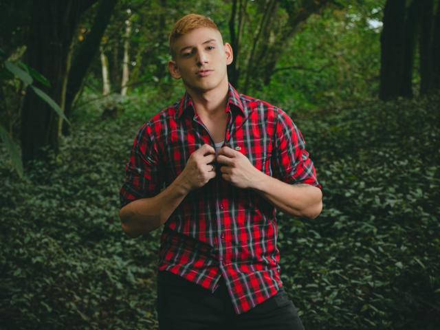 Jonathan Jeremiah, con una camicia a quadri rossa, pantaloni neri e capelli arancioni corti, tiene in mano i bottoni della camicia, è in piedi in una foresta, guarda leggermente di lato, è muscoloso, attraente e sicuro di sé..