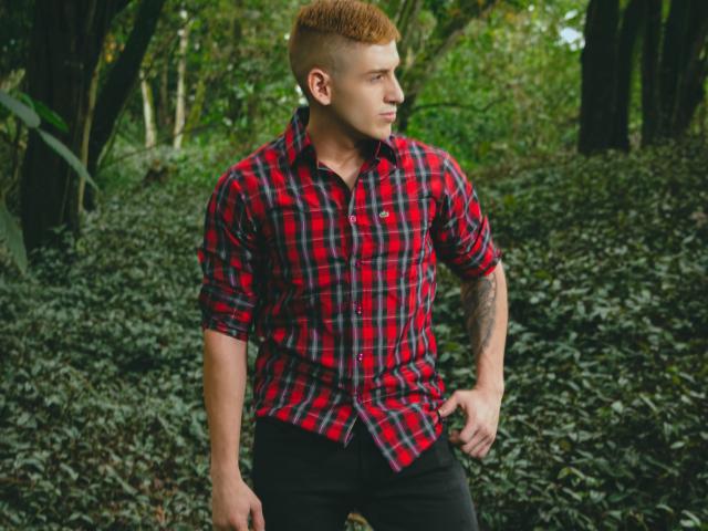 Jonathan Jeremiah, met een rood geruit shirt, zwarte broek en kort oranje haar, staat in het bos, met zijn rechterhand op zijn heup, en kijkt naar de zijkant. Hij heeft een tatoeage op zijn linkerarm..