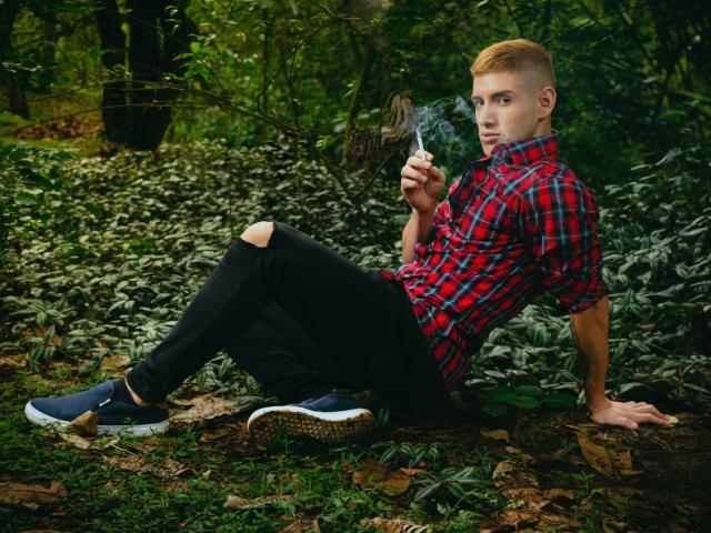 JonathanJeremiah with short, orange hair, red plaid shirt, black pants, and blue sneakers, smoking a cigarette, sitting on forest floor, legs crossed, looking at camera with a smug expression.
