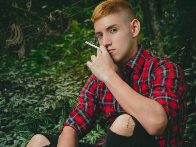JonathanJeremiah with short red hair, holding a cigarette to his nose, wearing a red plaid shirt and torn black jeans, sitting in a green forest.