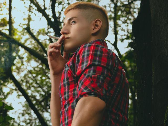 Jonathan Jeremiah, con cabello rojo corto, vistiendo una camisa de cuadros rojos, fumando un cigarrillo, sosteniéndolo cerca de sus labios, mirando hacia un lado con una expresión ligeramente seria..