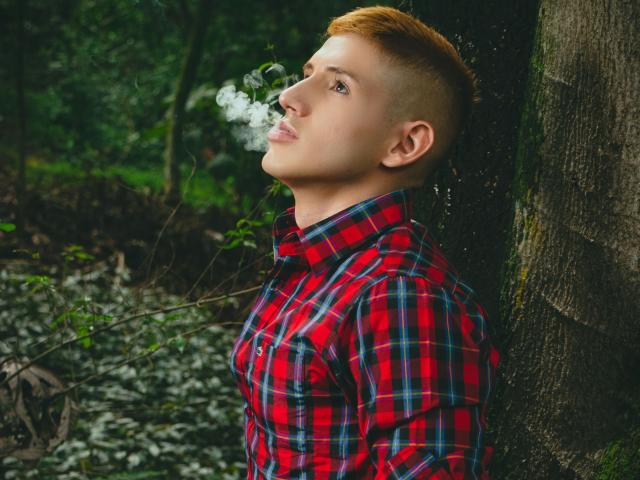 Jonathan Jeremiah, met kort rood haar, een rood geruit shirt, leunt tegen een boom, rookt, houdt zijn adem in, kijkt omhoog, bos op de achtergrond, broekspijp zichtbaar onder het shirt, groene begroeiing..