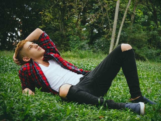Jonathan Jeremiah, met rood haar, ligt op het gras, gekleed in een rood geruit shirt, een wit t-shirt, zwarte jeans met gaten en blauwe schoenen, met zijn armen achter zijn hoofd, en ziet er ontspannen uit..