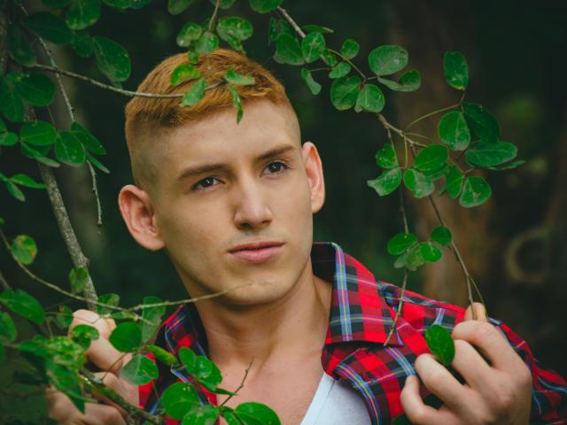 JonathanJeremiah with short red hair, blue eyes, and a clean-shaven face, wearing a red plaid shirt, holding green leaves, looking directly at the camera, with a serious expression.