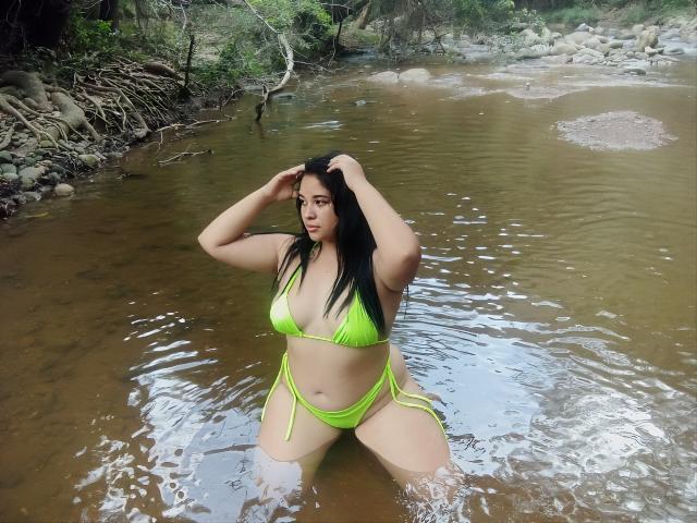 PamelaGoldwin, kneeling in a brown river, wearing a neon green bikini, hands in black hair, curvy with large tits, tan skin, and long black hair, looking at camera.