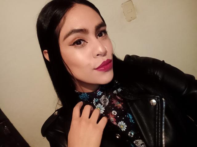 StellaBeckker with long black hair, wearing a black leather jacket, floral shirt, and pink lipstick, has her hand on her chest, looking at the camera with a slight smile.