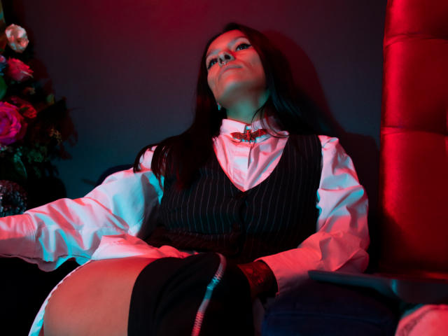 AmelieBlake with long dark hair, wearing a pinstriped vest over a white shirt, and a black skirt, sitting with her legs apart, revealing her bare, shiny, bare-ass. Red and blue light.