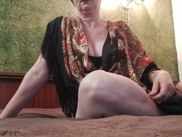 ShirlyNight has pale skin, small tits, and is wearing a colorful patterned shawl with black fringes, sitting on a brown bed with her leg raised, showing her ass.