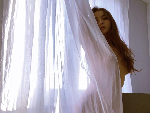 Dionisia with long brown hair, nude, standing by white curtains, holding them with her right arm, light from behind, soft shadows, gentle expression, subtle sensuality.