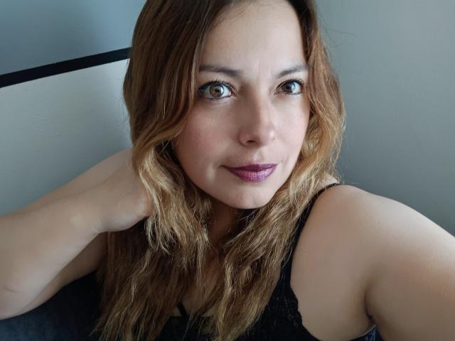 Seher has wavy brown hair, green eyes, and is wearing purple lipstick. She's in a black lace top, with her hand near her head, looking at the camera.