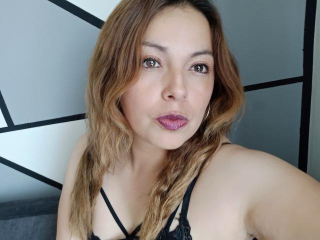 Seher with light brown wavy hair, wearing a black lace bra, pouting with pink lipstick, has brown eyes, and small hoop earrings, looking at the camera.