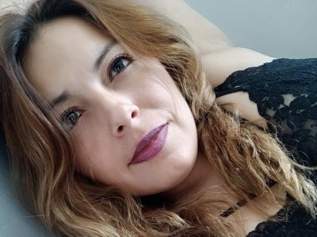 Seher's face, wavy brown hair, purple lipstick, black lace top, smiling, head resting on arm, gray eyes, fair skin, soft light, relaxed, cute, attractive, feminine.