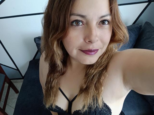 Seher with wavy brown hair, purple lipstick, black lace bra, smiling at the camera, showing her tits, and a cute face, taking a selfie from a high angle.